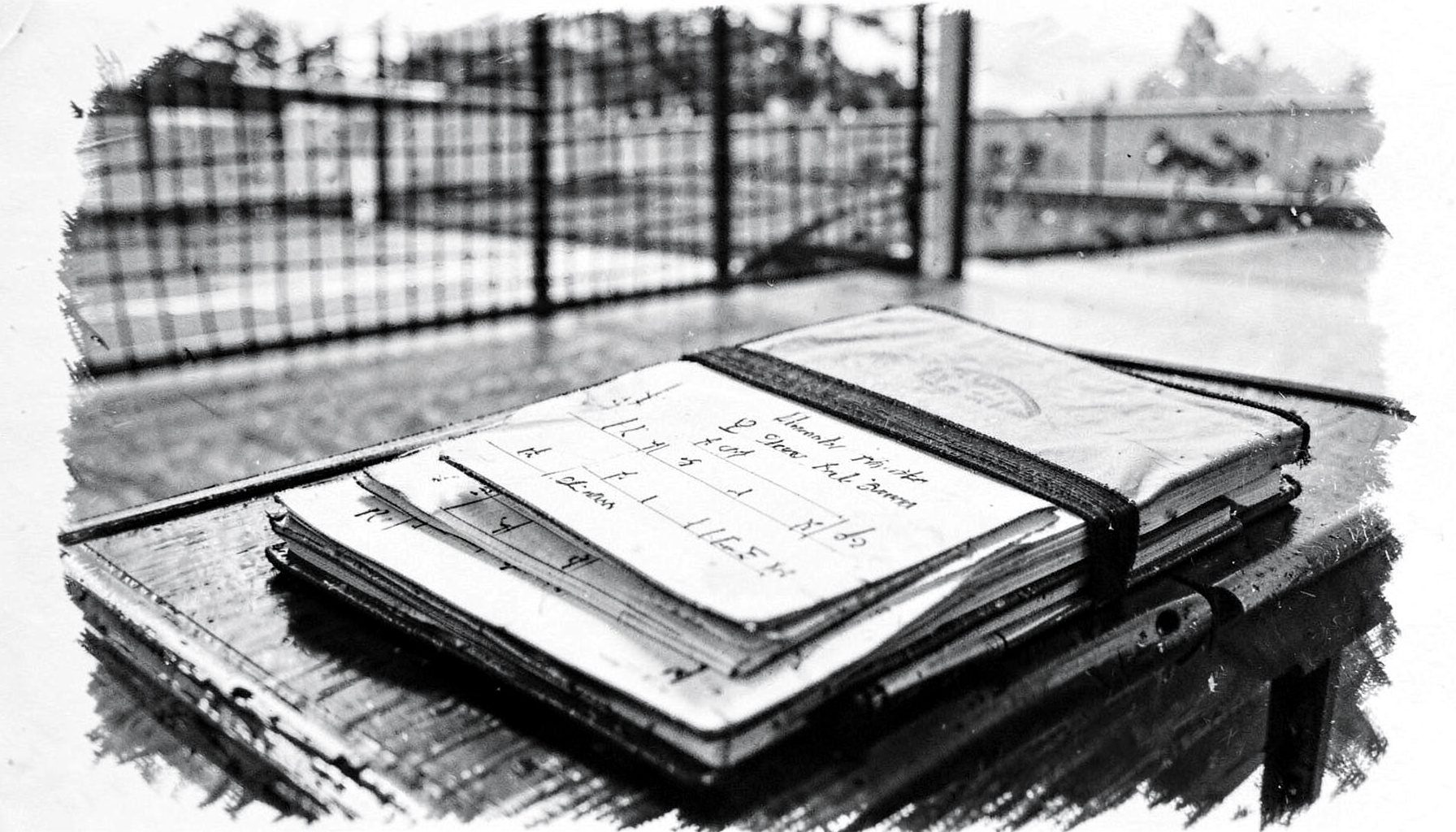 A charcoal-toned image of an official's scorebook and pen on a scorer's table, with the net and court visible in soft focus beyond